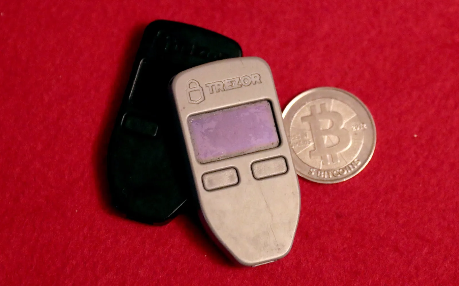Two Trezor One hardware cryptocurrency wallets and a physical Casascius five-bitcoin coin resting on red felt.